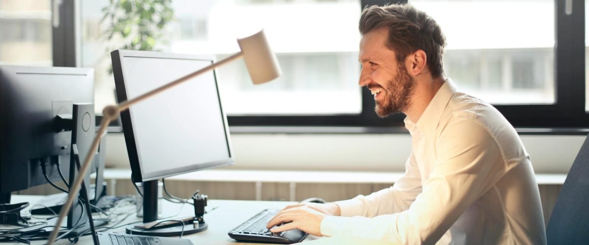 Man in White Dress Shirt Sitting on Black Rolling Chair While Facing Black Computer Set and Smiling